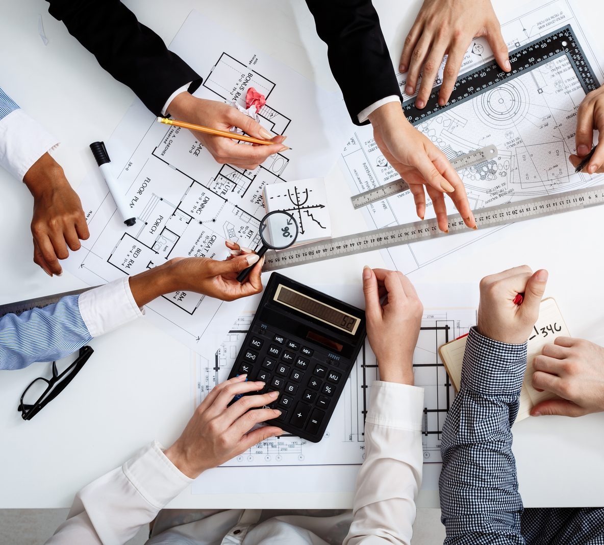 Overhead view of several people working over construction drawings with a calculator, scale rule and magnifying glass